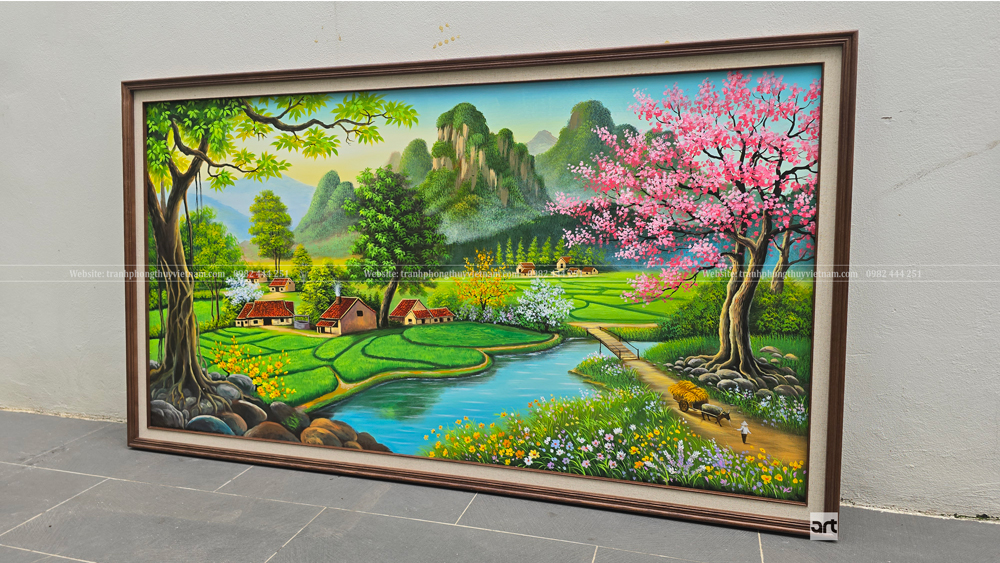 Tranh phong cảnh làng quê ven sông chụp nghiêng từ trái với ruộng xanh, mái ngói đỏ, núi đá và hoa sắc hồng Tranh phong cảnh làng quê ven sông chụp nghiêng từ trái với ruộng xanh, mái ngói đỏ, núi đá và hoa sắc hồng